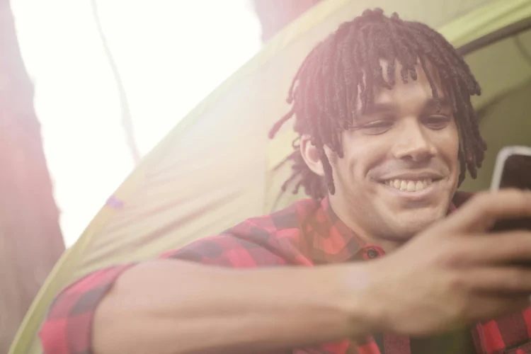 Young man smiling near tent, checking smartphone, representing relaxed lifestyle during Invisalign treatment duration.