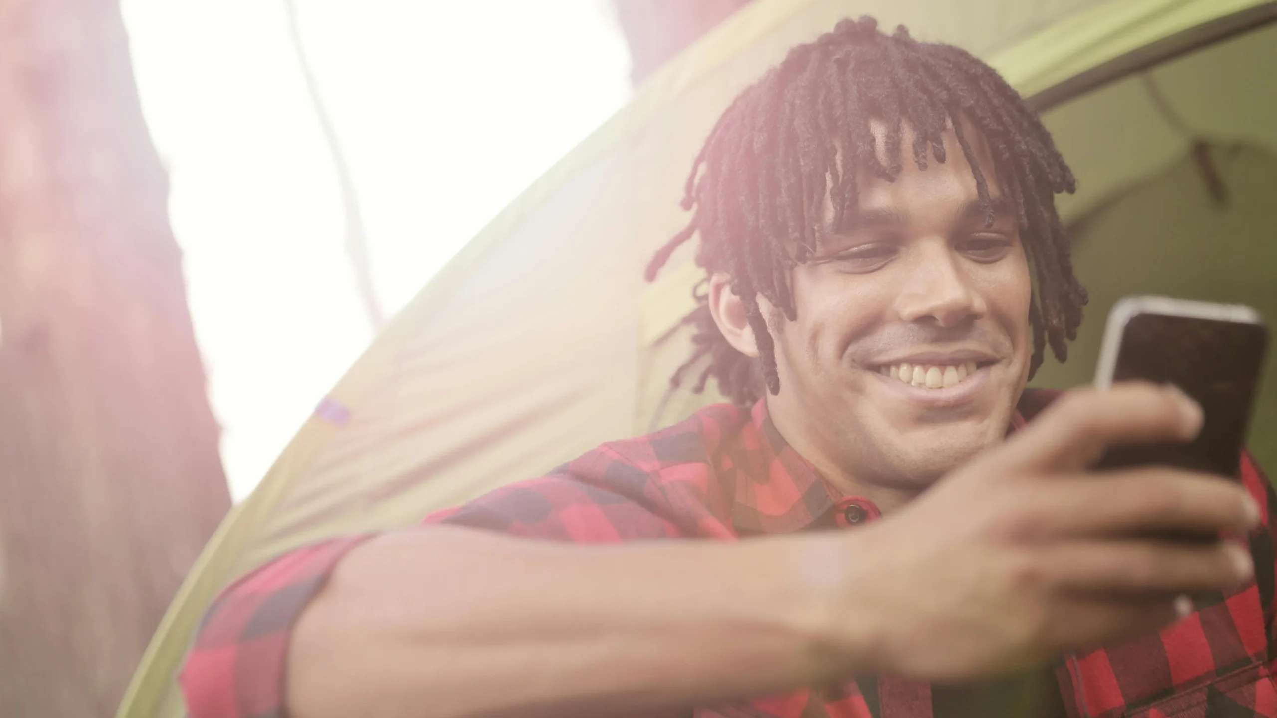 Young man smiling near tent, checking smartphone, representing relaxed lifestyle during Invisalign treatment duration.