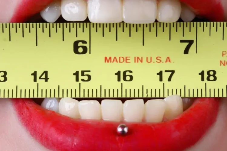 Mouth with red lipstick holding yellow tape measure, illustrating oral piercings dental issues.