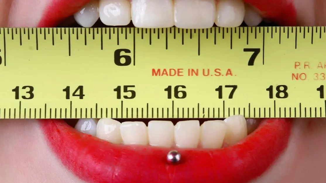 Mouth with red lipstick holding yellow tape measure, illustrating oral piercings dental issues.