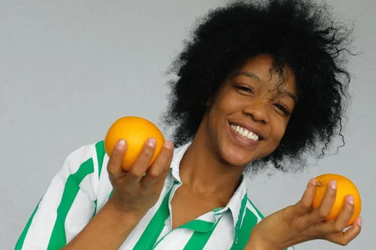 Cheerful woman with afro hair holding oranges, symbolizing ways to prevent and treat bad breath.