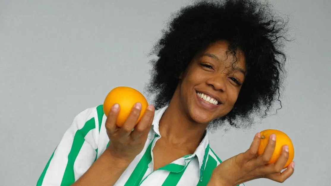 Cheerful woman with afro hair holding oranges, symbolizing ways to prevent and treat bad breath.